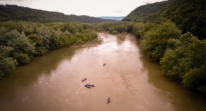 From above, a muddy body of water flows between tree-lined shores. There are small watercraft floating on the water. 